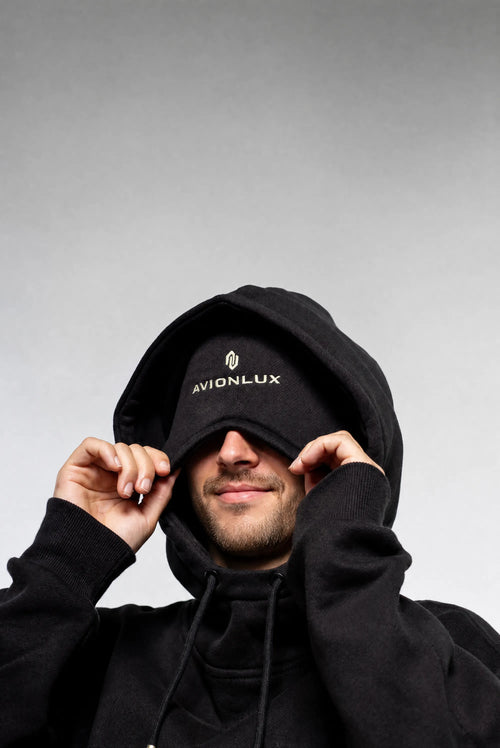 A man wearing a black hoodie and a black cap with the brand name 'AVIONLUX' embroidered in white on the front, holding the built-in eye mask over his eyes against a gray studio background.