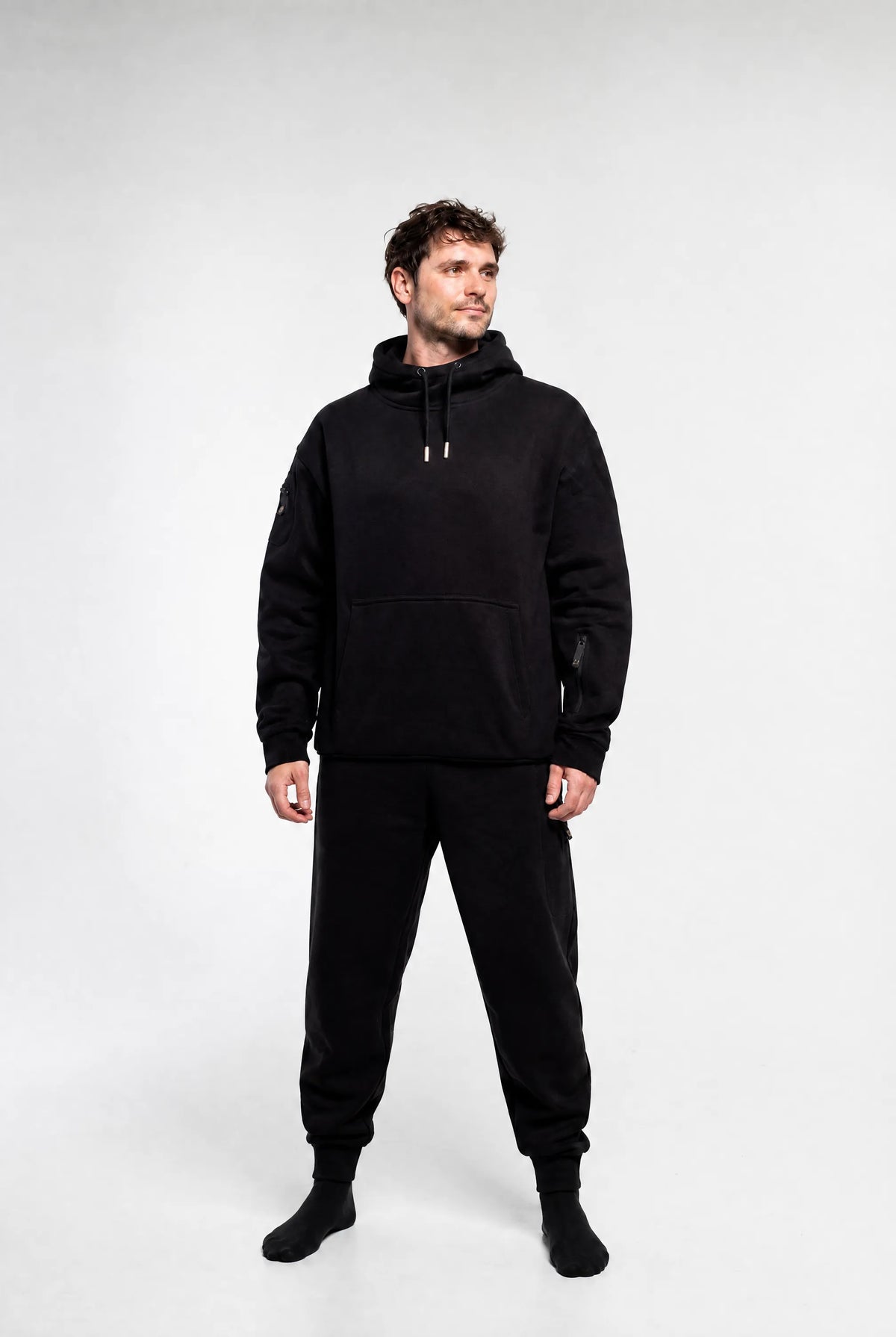 A man modeling a black pullover hoodie and matching black jogger pants against a light gray studio background.