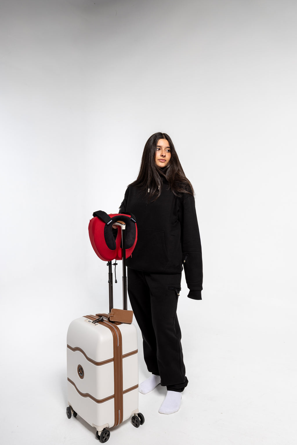 A woman in a black sweatshirt and pants stands next to a cream-colored hard-shell suitcase with brown trim and a red and black travel neck pillow, set against a plain light gray studio background.