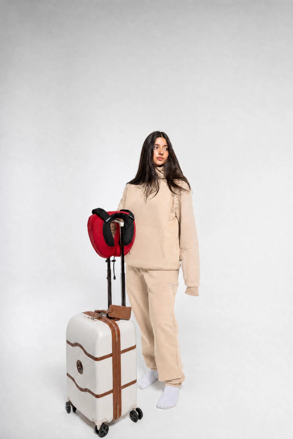 A woman stands against a light gray studio background wearing a beige tracksuit and white socks, holding a cream hard-shell suitcase with brown trim and a red backpack resting on top.