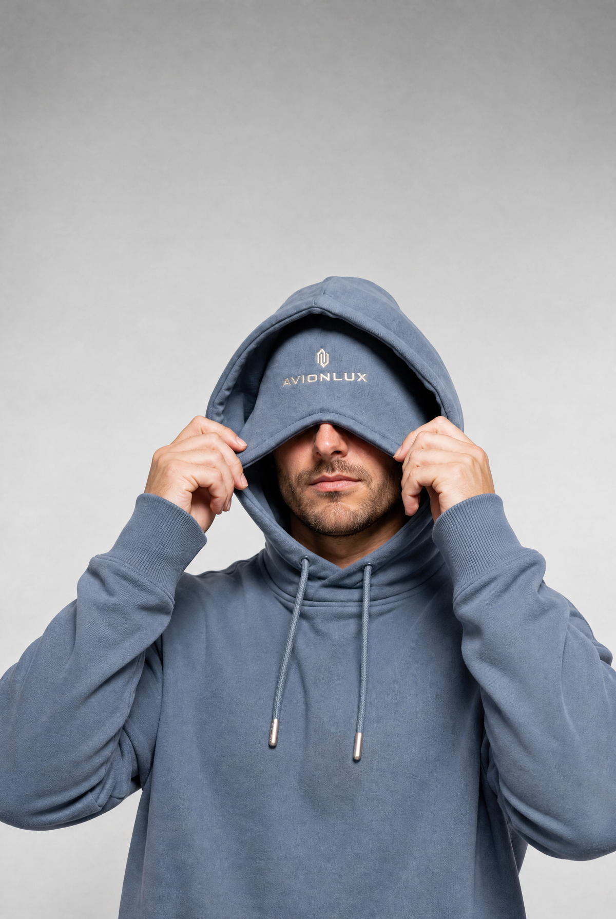 A man wearing a slate blue hoodie with the brand name 'AVIONLUX' embroidered in white on the chest, holding the built-in eye mask up over his eyes against a neutral gray background.
